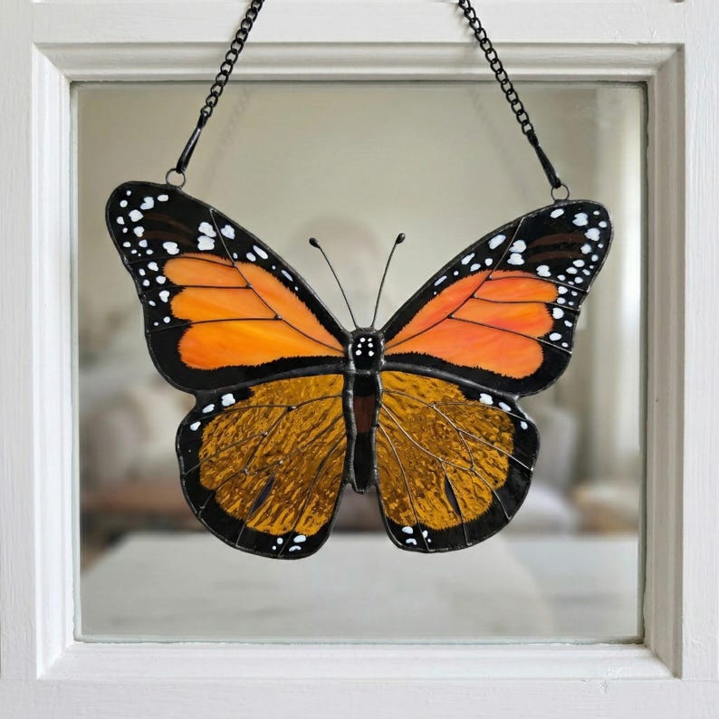 K&ouml;nnte beinhalten: Glasmalerei Monarchfalter mit orangefarbenen und braunen Fl&uuml;geln, schwarzen Umrissen und wei&szlig;en Punkten. Der Schmetterling wird von einer schwarzen Kette gehalten und in einem wei&szlig; gerahmten Fenster ausgestellt. Dekoratives Kunstwerk.