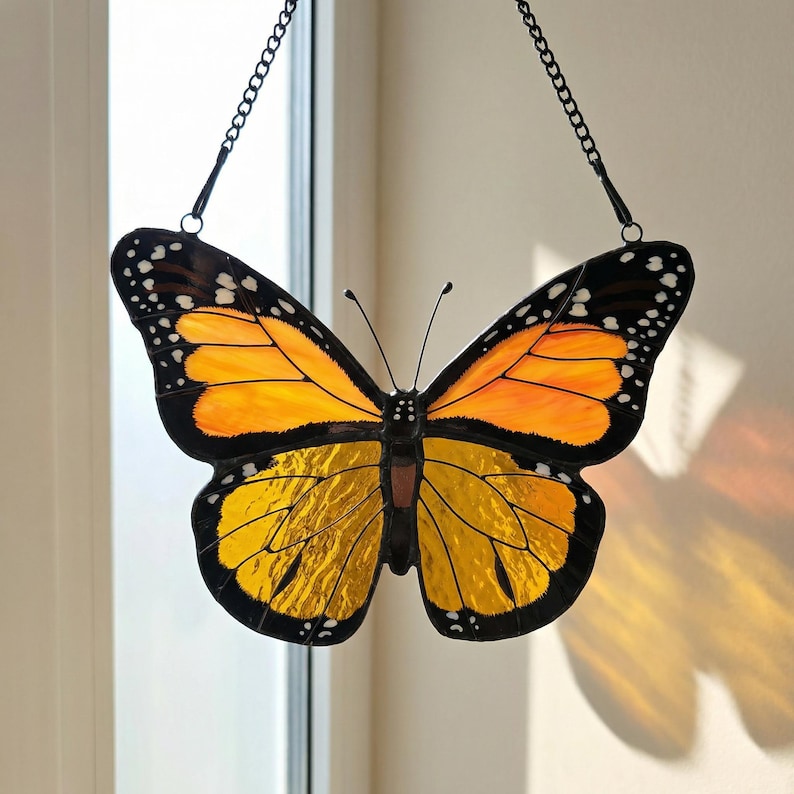 K&ouml;nnte beinhalten: Buntglas-Schmetterling-Suncatcher mit orangefarbenen und gelben Fl&uuml;geln, schwarzen R&auml;ndern und wei&szlig;en Punkten. Der dekorative Schmetterling wird von einer schwarzen Kette gehalten, ideal f&uuml;r Fenster- oder Wanddekoration.