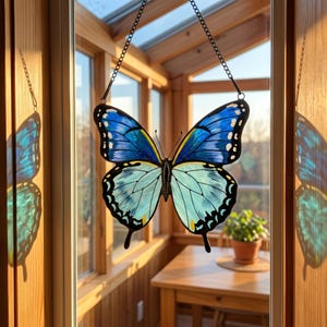 May include: A stained glass butterfly suncatcher with blue and light blue wings, black outlines, and yellow accents. The butterfly is suspended by a chain in front of a window, with a wooden interior visible in the background.
