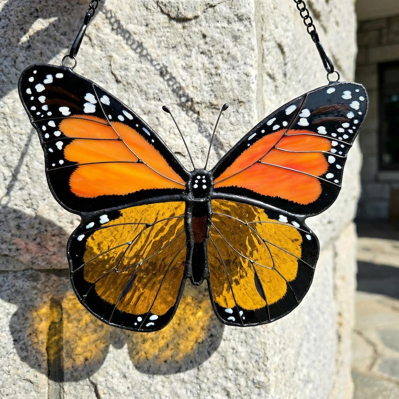 K&ouml;nnte beinhalten: Ein Buntglas-Schmetterling mit orangefarbenen und gelben Fl&uuml;geln, schwarz umrandet. Der Schmetterling hat wei&szlig;e Punkte an den Fl&uuml;gelkanten und wird von einer schwarzen Kette gehalten. Die Fl&uuml;gel sind symmetrisch und detailliert.