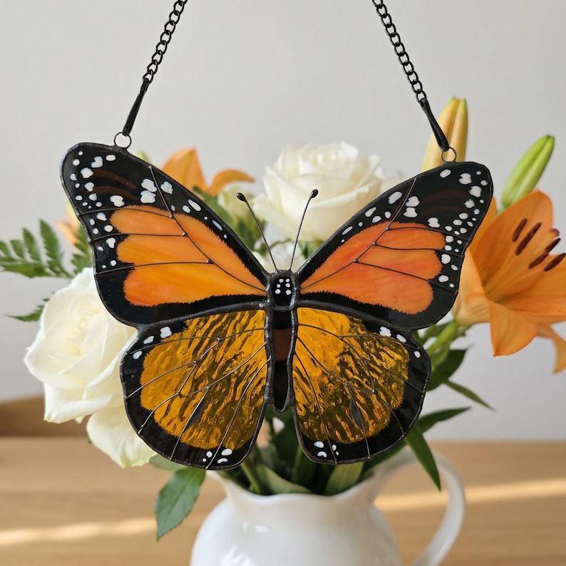 K&ouml;nnte beinhalten: Ein Buntglas-Schmetterling mit orangefarbenen und schwarzen Fl&uuml;geln, verziert mit wei&szlig;en Punkten. Der Schmetterling h&auml;ngt an einer schwarzen Kette. Im Hintergrund wei&szlig;e Rosen und orangefarbene Lilien in einer wei&szlig;en Vase, die eine lebendige Darstellung bilden.