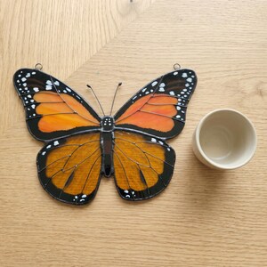 K&ouml;nnte beinhalten: Ein Buntglas-Schmetterling mit orangefarbenen und schwarzen Fl&uuml;geln, wei&szlig;en Punkten und einer schwarzen Umrandung. Der Schmetterling befindet sich neben einer kleinen, cremefarbenen Keramikschale. Der Hintergrund ist eine helle Holzoberfl&auml;che.