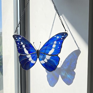 May include: A stained glass butterfly with vibrant blue wings and white accents, suspended by a black chain. The butterfly's wings are detailed with white spots and patterns. The piece is hanging in front of a window.