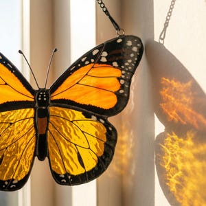 K&ouml;nnte beinhalten: Ein Glasfenster-Schmetterling-Sonnenf&auml;nger mit orangefarbenen, gelben und schwarzen Details. Der Schmetterling hat ein detailliertes Fl&uuml;gelmuster und wird von einer Kette gehalten. Der Sonnenf&auml;nger wirft einen farbenfrohen Schatten.