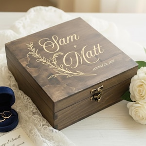 May include: A wooden box with the names "Sam + Matt" and the date "August 22, 2026" engraved on the lid. A blue velvet ring box with two gold rings is open next to the wooden box. A bouquet of white roses is also present.