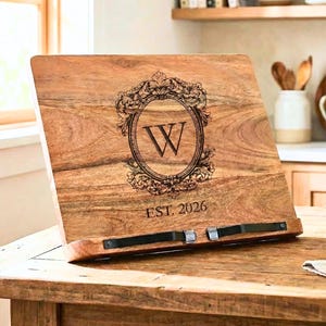 May include: Wooden cookbook stand with a decorative oval design featuring the letter "W" and the text "EST. 2026". The stand is made of light brown wood and has a metal bar to hold the book open. The stand is on a wooden table.