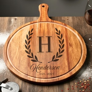 May include: A round wooden cutting board with a handle, engraved with the letter "H" inside a laurel wreath, the name "Henderson", and the year "EST. 2025". The board is surrounded by a circular groove.
