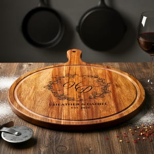 May include: A wooden pizza serving board with a handle, engraved with initials and a date, sits on a wooden table. A glass of red wine, a small bowl of spices, and a pizza cutter are also on the table. Two black pans hang in the background.