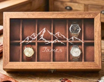 May include: Wooden watch display case with a glass top, featuring a mountain design and the name "James." The case holds several watches, with three visible. The case is made of wood and has a brown interior.