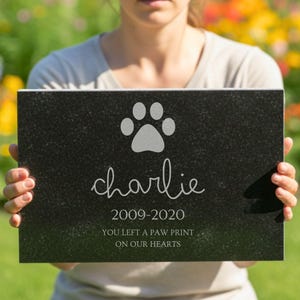 K&ouml;nnte beinhalten: Eine rechteckige Gedenktafel aus schwarzem Granit f&uuml;r Haustiere mit einer eingravierten Pfotenabdruck-Grafik, dem Namen Charlie, den Jahren 2009-2020 und dem Text YOU LEFT A PAW PRINT ON OUR HEARTS. Der Stein hat eine polierte, leicht gesprenkelte Oberfl&auml;che.
