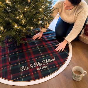 May include: A round Christmas tree skirt featuring a dark green, red, and navy blue plaid pattern with a white border. The fabric is personalized with the white script text "Mr & Mrs Holland" and the block text "EST 2026" printed on the surface.