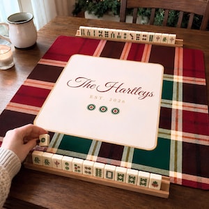 May include: A square Mahjong mat with a red, green, and white plaid pattern featuring a central white square printed with the text The Hartleys EST. 2026 and three circular icons. Two wooden tile racks hold ivory Mahjong tiles with green and red symbols.