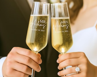 May include: Two champagne flutes filled with a golden liquid. Each glass is engraved with "Mr. Adams" or "Mrs. Adams" and the date "October 18, 2025". The flutes are held up in a celebratory toast.