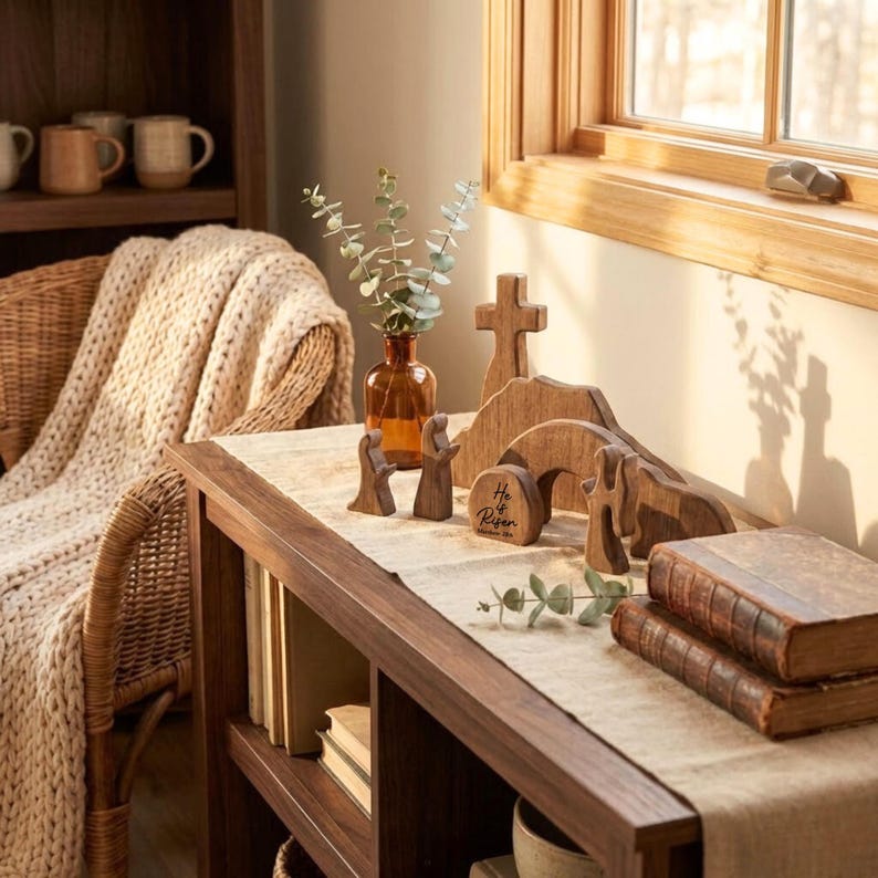 May include: Wooden Easter decorations on a wooden table. The decorations include a cross, a tomb with the words "He is Risen", and figures of people. A vase with greenery and two old books are also on the table. A woven chair and a wooden shelf are in the background.