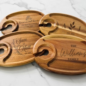 May include: Wooden wine serving trays with a circular cutout for a wine glass and a groove for snacks. Each tray is engraved with personalized text, including names, dates, and family names. The trays are made of light brown wood.