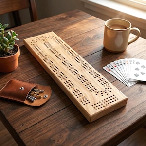 May include: A wooden cribbage board with multiple rows of holes for pegs, alongside a leather pouch with pegs, a deck of cards, and a mug of coffee. The board is made of light-colored wood.