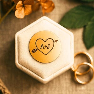 May include: A gold-colored, round disc with a heart and arrow design, engraved with the letters "A+J", rests on a beige hexagonal ring box. Two gold wedding bands are visible in the background. The image has a soft, romantic aesthetic.