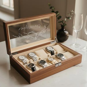 May include: A wooden watch box with a glass top displaying eight watches. The interior has beige cushions for each watch. The glass top is etched with the name "Christopher" and a mountain range design.