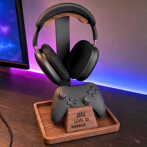 May include: A dark wood and black metal headphone and controller stand featuring a square base with a tray. The front of the wooden base is engraved with the text JAKE LEVEL 30 and a loading bar graphic. A pair of black over-ear headphones rests on top.