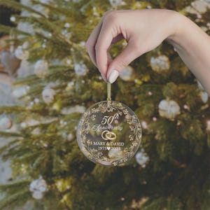May include: A clear glass Christmas ornament with gold text and accents. The ornament reads "50th Anniversary" with the names "Mary & David" and the years "1975 2025". The ornament is held by a hand in front of a Christmas tree.