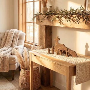 May include: Wooden Easter scene with a cross, tomb, and figures. The scene is on a wooden table with a cream-colored runner. The table is in a room with a window and a beige chair with a knitted throw.