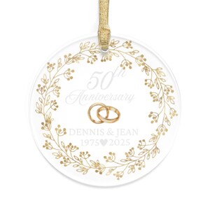 May include: A clear, round ornament with gold accents, featuring the text "50th Anniversary" and the names "Dennis & Jean" with the years 1975 and 2025. The design includes a gold floral wreath and two gold wedding rings.