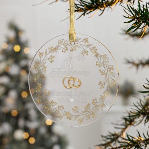 May include: A clear, round ornament with gold text and accents. The ornament features the words "50th Anniversary", two gold wedding rings, and the names "DENNIS & JEAN 1975 2025". A gold ribbon is attached for hanging.