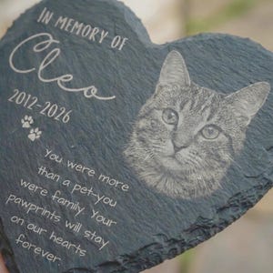 May include: Heart-shaped slate pet memorial with a detailed engraved portrait of a cat. The memorial includes the name "Cleo", dates 2012-2026, paw prints, and a heartfelt message. The slate is dark gray.