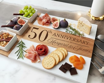 May include: A wooden and marble serving board with the text "50th Anniversary Margaret & James est. 1975". The board is filled with grapes, figs, nuts, cheese, crackers, prosciutto, chocolate, and apricots. Serving utensils are on the side.