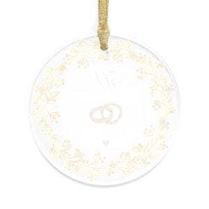 May include: Clear, round ornament with a gold floral wreath design. The ornament features two intertwined rings and text. A gold ribbon is attached for hanging. Ideal for weddings or anniversaries.
