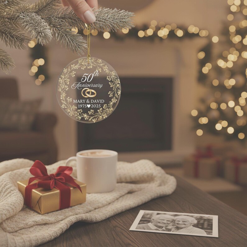 May include: A clear, round ornament with gold text and accents, celebrating a 50th anniversary. The ornament features the text "50th Anniversary", two wedding rings, and the names "Mary & David 1975 2025". The ornament is held by a hand.