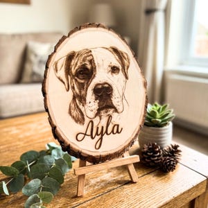 May include: A wooden plaque with a dog portrait and the name "Ayla" engraved on it. The dog is a Boxer or similar breed, with a brown and white coat. The plaque is displayed on a small wooden easel.