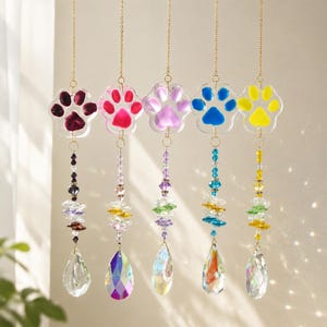 May include: Five hanging suncatchers featuring clear acrylic paw prints in dark red, bright pink, light purple, blue, and yellow. Each paw print is suspended by a gold-toned chain with multi-colored crystal beads and a clear teardrop prism.