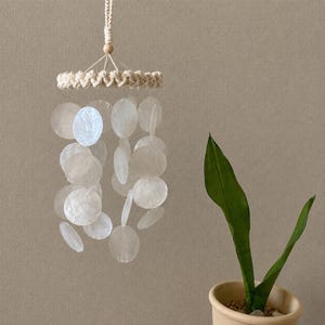 May include: A hanging mobile featuring a circular macrame frame in off-white cord with a central wooden bead. Multiple strands of translucent, circular capiz shell discs hang from the frame, creating a layered, shimmering effect in white.