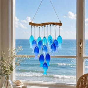 K&ouml;nnte beinhalten: Ein h&auml;ngendes Mobile mit einem horizontalen St&uuml;ck Treibholz, an dem mehrere blaue Fischformen aus Glas an d&uuml;nnen Schn&uuml;ren befestigt sind. Die Fische variieren in Blaut&ouml;nen von hell bis tief Kobalt und sind in einem kaskadierenden Muster angeordnet.