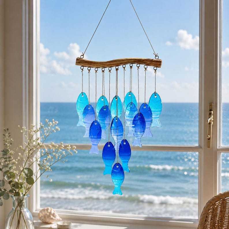 K&ouml;nnte beinhalten: Ein h&auml;ngendes Mobile mit einem horizontalen St&uuml;ck Treibholz, an dem mehrere blaue Fischformen aus Glas an d&uuml;nnen Schn&uuml;ren befestigt sind. Die Fische variieren in Blaut&ouml;nen von hell bis tief Kobalt und sind in einem kaskadierenden Muster angeordnet.