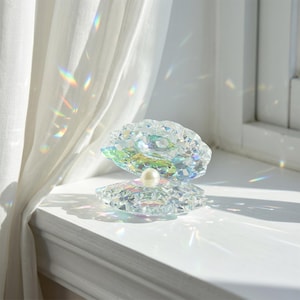 Puede incluir: Un adorno de cristal transparente facetado con forma de concha abierta, con una perla blanca única, redonda y lisa, situada en el centro. La superficie de cristal refleja la luz, creando patrones prismáticos de arcoíris en las superficies.