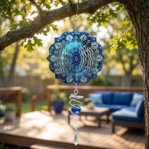 May include: A blue and silver metal wind spinner with a mandala design hangs from a tree branch. The spinner has a spiral tail with a blue glass ball and a crystal at the end. The background shows a deck and outdoor furniture.