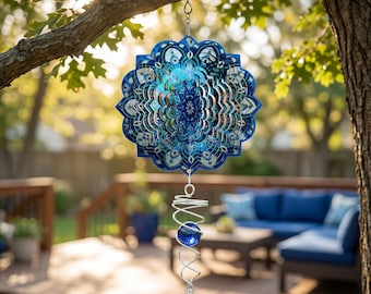 May include: A blue and silver metal wind spinner with a mandala design hangs from a tree branch. The spinner has a spiral tail with a blue glass ball and a crystal at the end. The background shows a deck and outdoor furniture.