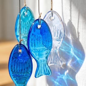K&ouml;nnte beinhalten: Eine Sammlung von vier durchscheinenden Fischornamenten aus Glas in verschiedenen Blaut&ouml;nen, die von hellem Himmelblau bis zu tiefem Kobaltblau reichen. Jeder Fisch hat ein geformtes Rippenmuster und ein kleines Loch zum Aufh&auml;ngen.
