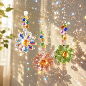 Op de afbeelding: Drie hangende kristallen bloem zonnevangers. Elke bloem heeft een andere kleur, waaronder helder, roze en groen. De bloemen zijn opgehangen aan een zilveren ketting en versierd met kleurrijke kralen. Het zonlicht creëert een sprankelend effect.