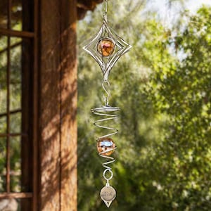 May include: A silver metal wind spinner with a diamond-shaped top and a spiral design. It features two orange glass orbs and a pendant that reads "Tish". This decorative item is designed for outdoor use and hangs from a hook.
