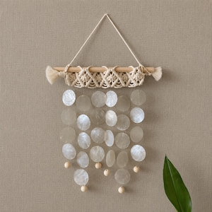 May include: A wall hanging featuring a horizontal wooden dowel with cream-colored macrame cord knotted in a diamond pattern. Suspended from the macrame are multiple translucent, circular capiz shell discs arranged in rows, accented with small wooden beads.
