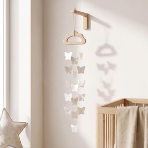 Puede incluir: Un móvil colgante con múltiples adornos blancos en forma de mariposa suspendidos de un soporte de madera clara con forma de nube. El soporte está fijado a un brazo de madera montado en la pared. Las mariposas tienen un acabado nacarado.