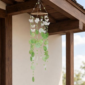 May include: A hanging mobile featuring a circular wooden frame with multiple strands of translucent green circular discs and white butterfly-shaped ornaments. The strands are accented with small green beads and suspended by thin white strings.
