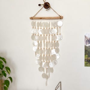 May include: A wall hanging featuring multiple strands of circular, iridescent white shell discs interspersed with small, round, light-coloured wooden beads. The strands are suspended from a horizontal piece of light brown driftwood.