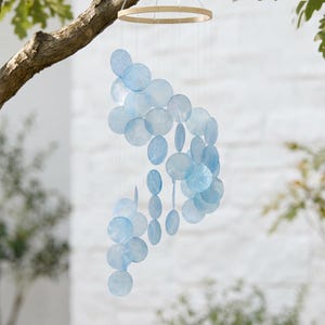 May include: A hanging mobile featuring numerous translucent, light blue circular discs suspended from a circular wooden frame by thin, clear strings. The discs are arranged in a cascading, vertical pattern, creating a layered, airy appearance.