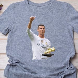 May include: Gray t-shirt featuring a graphic of a soccer player in a white jersey, holding a golden shoe trophy. The player is raising his arm in a celebratory gesture. The jersey has the Emirates logo.