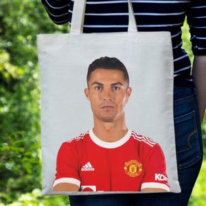 May include: White tote bag featuring a portrait of a soccer player in a red jersey with white stripes and the Adidas logo. The jersey also displays the Manchester United emblem. The bag is held against a green background.