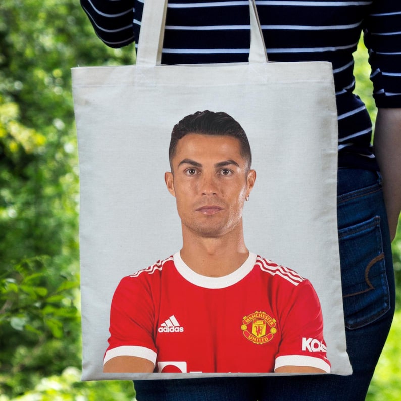 May include: White tote bag featuring a portrait of a soccer player in a red jersey with white stripes and the Adidas logo. The jersey also displays the Manchester United emblem. The bag is held against a green background.